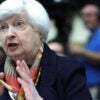 Treasury Secretary Janet Yellen Testifies at a Hearing in a black jacket with a scarf. Sitting at a microphone and gesturing with her hands