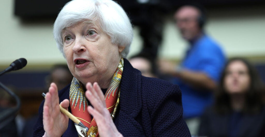 Treasury Secretary Janet Yellen Testifies at a Hearing in a black jacket with a scarf. Sitting at a microphone and gesturing with her hands