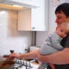 A father with an infant in his arms stirring a pot on a gas stove in the kitchen