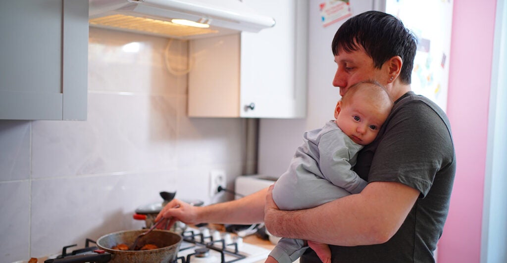 A father with an infant in his arms stirring a pot on a gas stove in the kitchen