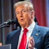 Donald Trump in a blue suit and red tie with a flag in the background speaks at a microphone