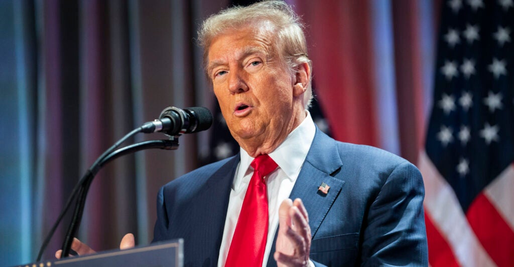 Donald Trump in a blue suit and red tie with a flag in the background speaks at a microphone