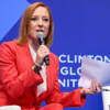 MSNBC host Jen Psaki in a red suit with a microphone in her hand sitting in a chair on stage with a blue background