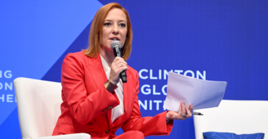 MSNBC host Jen Psaki in a red suit with a microphone in her hand sitting in a chair on stage with a blue background