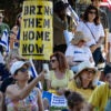 People sitting and standing on the grass with a big "Bring Them Home Now" sign posted on a tree.