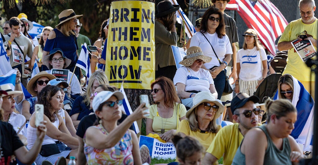 People sitting and standing on the grass with a big "Bring Them Home Now" sign posted on a tree.