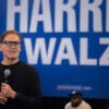 Mark Cuban, standing in front of the Harris-Walz sign, holds a mic in his hand as he answers questions from people in the audience.