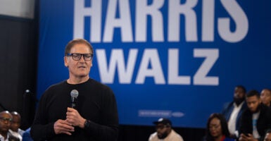 Mark Cuban, standing in front of the Harris-Walz sign, holds a mic in his hand as he answers questions from people in the audience.