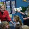 Hillary Clinton sits in front of a crowd of pre-K students, dressed in a red blazer, speaking to them during a reading of "The Very Hungry Caterpillar."