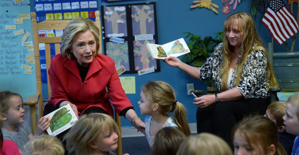 Hillary Clinton sits in front of a crowd of pre-K students, dressed in a red blazer, speaking to them during a reading of "The Very Hungry Caterpillar."