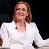 Jen Psaki, in a white suit, sits on a panel, smirking and shrugging her shoulders.