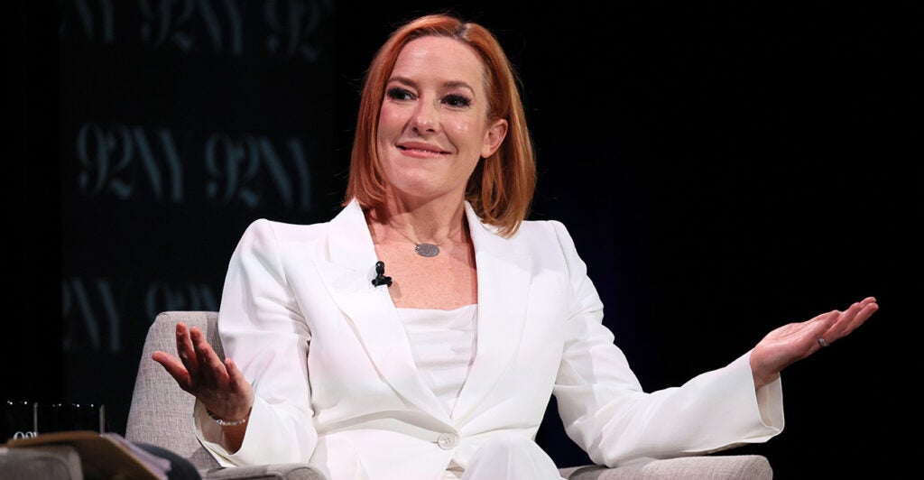 Jen Psaki, in a white suit, sits on a panel, smirking and shrugging her shoulders.