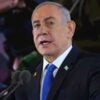 Benjamin Netanyahu in a dark suit and blue tie speaking at a microphone