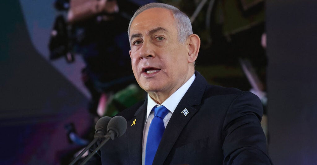 Benjamin Netanyahu in a dark suit and blue tie speaking at a microphone