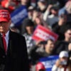 President Donald Trump, in a suit, black overcoat, and red Make America Great Again baseball cap stands outdoors at a rally with a crowd of rallygoers in back of him and his arm raised, pointing to the crowd in front of him.