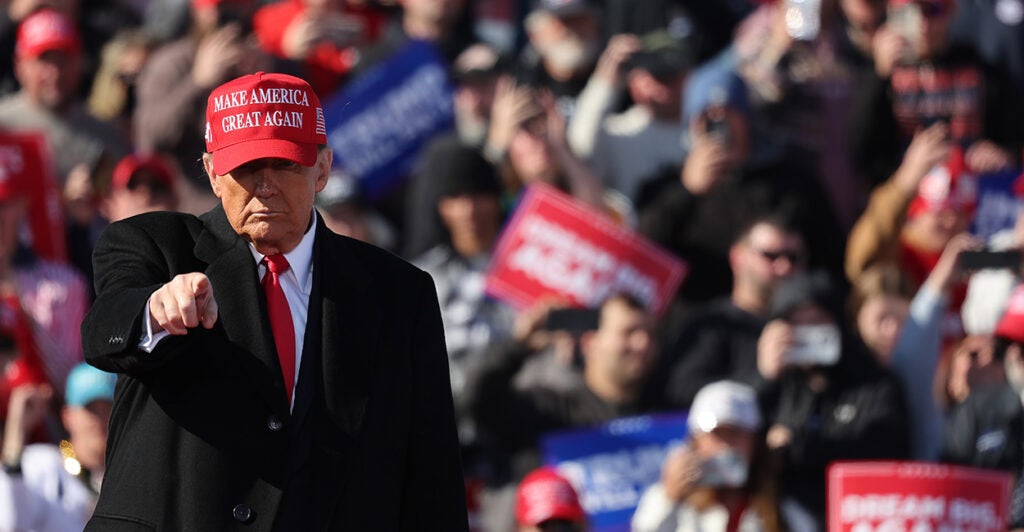 President Donald Trump, in a suit, black overcoat, and red Make America Great Again baseball cap stands outdoors at a rally with a crowd of rallygoers in back of him and his arm raised, pointing to the crowd in front of him.