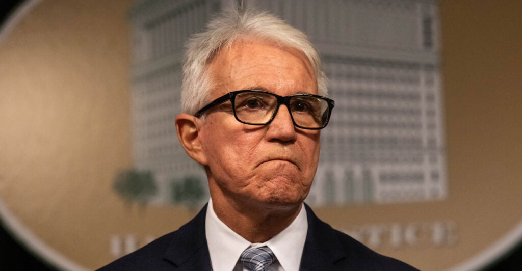 Los Angeles County District Attorney George Gascón in a dark suit and glasses with a frown talks to the media at a news conference