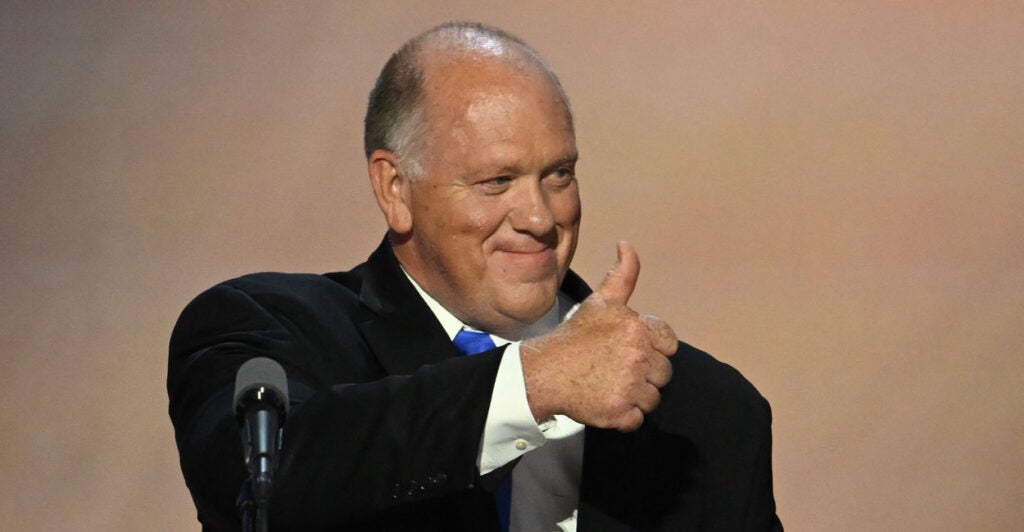 Tom Homan on stage at a lectern smiling and giving a thumbs-up to the audience.