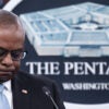 US Secretary of Defense Lloyd Austin III, in a suit, hangs his head and stands before a sign. that reads "The Pentagon."