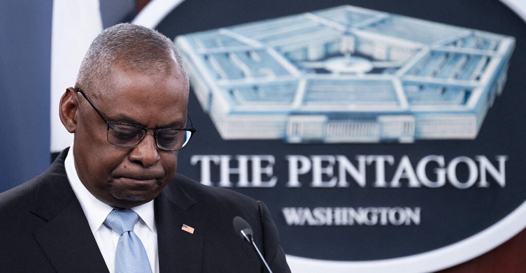 US Secretary of Defense Lloyd Austin III, in a suit, hangs his head and stands before a sign. that reads "The Pentagon."