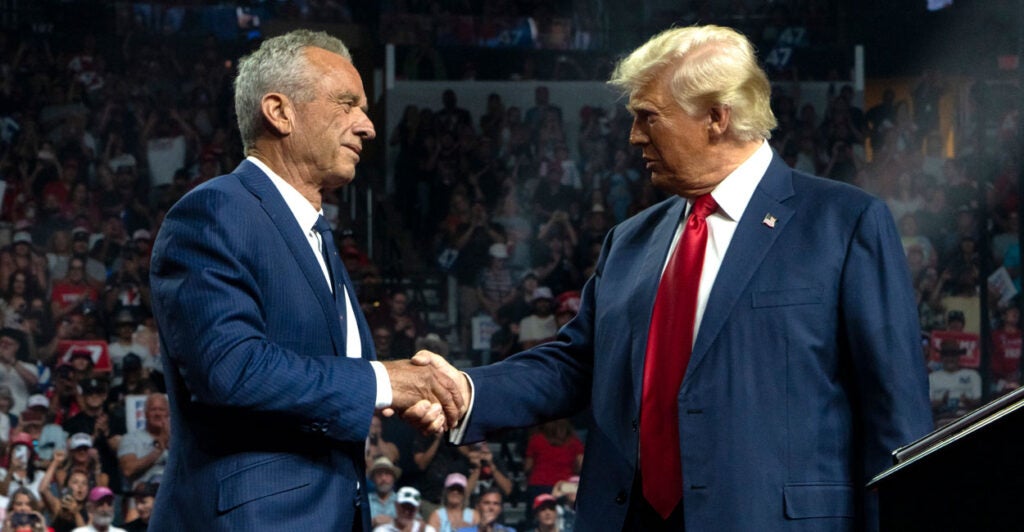 Robert F. Kennedy, Jr. and former President Donald Trump, both in dark blue suits, shake hands on stage at a Trump rally