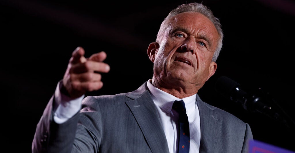 Robert F Kennedy Junior in a gray suit and a dark tie In front of a black background gestures by raising his arm and pointing his finger.