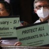 two older people, a man and a woman, people hold up signs saying “sanctuary” and “protect all immigrants in LA”