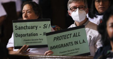 two older people, a man and a woman, people hold up signs saying “sanctuary” and “protect all immigrants in LA”