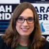 Candidate Sarah McBride and long hair and glasses Is in front of a wall of McBride's campaign signs