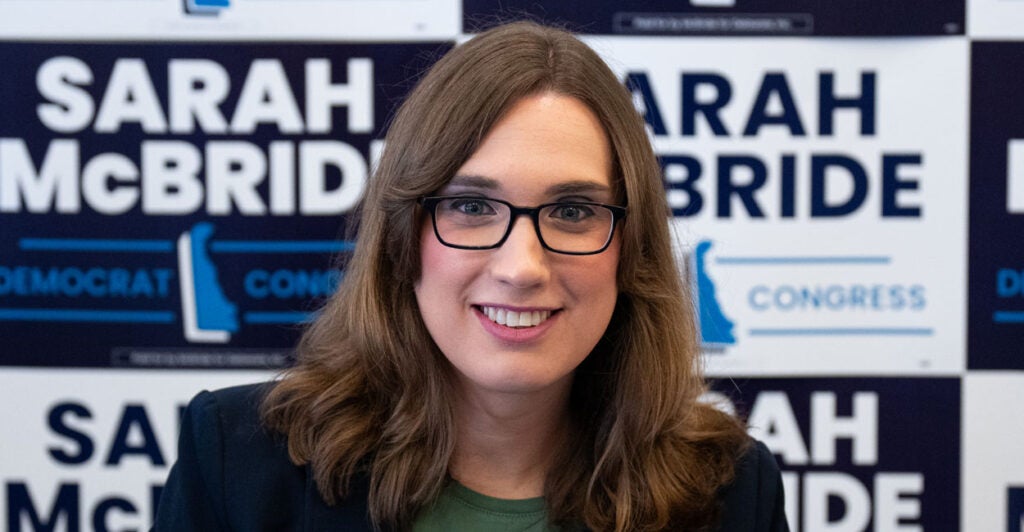 Candidate Sarah McBride and long hair and glasses Is in front of a wall of McBride's campaign signs