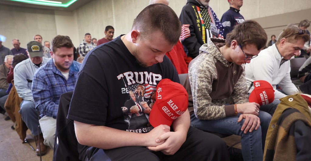 Man in Trump shirt holds a MAGA hat to his chest as he prays at a rally for President-elect Donald Trump.