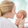 A new mother lays back in her hospital bed shortly after childbirth, with her baby held out in front of her as they bond with one another.