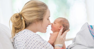 A new mother lays back in her hospital bed shortly after childbirth, with her baby held out in front of her as they bond with one another.