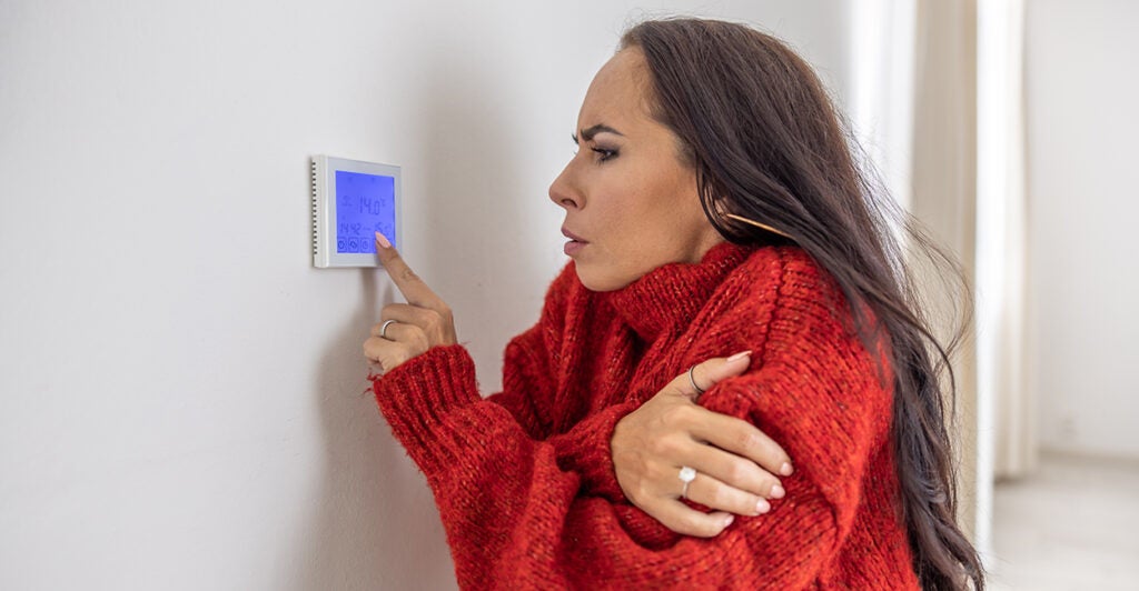 A cold woman at home wearing red sweater raises the temperature on a thermostat.