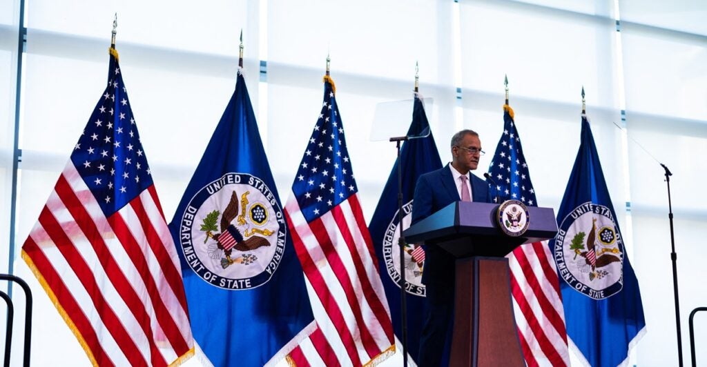 Richard Verma, deputy secretary of State for Management and Resources, stands at a podium with several flags behind him.