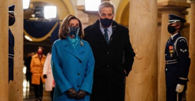 Nancy Pelosi wears a blue coat and black mask as she stands next to her husband, Paul Pelosi, who wears a black coat and black mask.