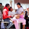 A Haitian woman with curly hair sits with her large pregnant belly protruding from her striped shirt and pink pants as her partner in a bright orange sweatshirt holds her closely.