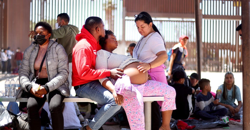 A Haitian woman with curly hair sits with her large pregnant belly protruding from her striped shirt and pink pants as her partner in a bright orange sweatshirt holds her closely.