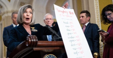 Sen. Joni Ernst dressed in a black blazer with her hair down, holds up a long list titled "Naughty List" in red ink, which includes all of the federal agencies with low percentages of staff who work in the office.