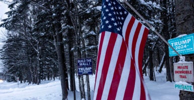 Along a wintery highway, an American flag hangs nailed to a snow-covered tree among pro-Trump campaign signs supporting the building of a border wall.