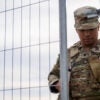 A black National Guard soldier dressed in a camouflage uniform pulls a silver-wired security gate closed.