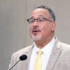 Miguel Cardona, wearing a tan suit jacket coat and yellow necktie, speaks during a commemoration ceremony.