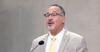 Miguel Cardona, wearing a tan suit jacket coat and yellow necktie, speaks during a commemoration ceremony.