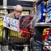 An older man wearing dark-rimmed glasses looks at a coupon book as he pushes a black grocery cart.