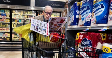 An older man wearing dark-rimmed glasses looks at a coupon book as he pushes a black grocery cart.