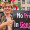 A smiling trangender activist wearing a black vest and colorful jester hat holds a sign that reads, "No Pride In Genocide."