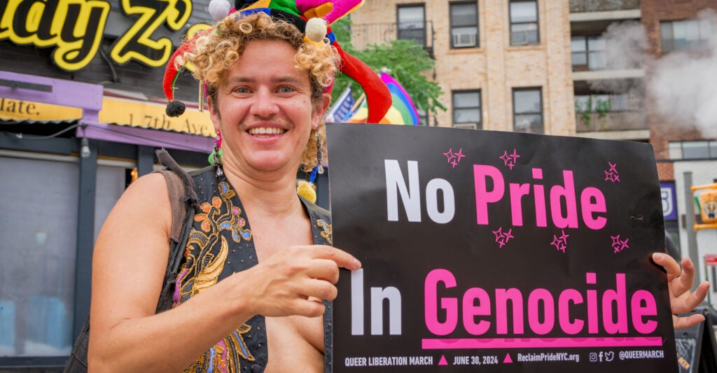 A smiling trangender activist wearing a black vest and colorful jester hat holds a sign that reads, "No Pride In Genocide."