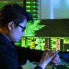 An Asian hacker wearing glasses and a black sweatshirt sits in a dark room while using a smartphone and coding on multiple monitors displaying cybersecurity-related data.