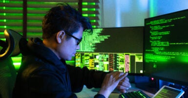 An Asian hacker wearing glasses and a black sweatshirt sits in a dark room while using a smartphone and coding on multiple monitors displaying cybersecurity-related data.