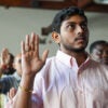 A young man with black hair and brown skin wears a light pink shirt as he raises his right hand with a gold ring on it during his naturalization ceremony.